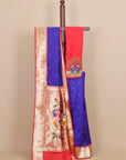 A royal blue and red saree draped over a wooden hanger against a beige background. The saree features a wide gold zari border with floral embroidery, a red pallu with peacock motifs, and small gold dots on the blue body.