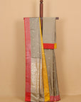 A traditional Indian saree draped over a wooden hanger against a solid beige background. The saree has a gray body with small white motifs, a red border, a yellow border, and a light gray pallu with a red and yellow border.