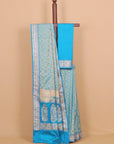 A light blue and turquoise silk saree with intricate silver zari motifs, draped over a wooden hanger against a solid beige background.