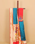 A traditional Indian silk saree draped over a wooden hanger against a solid beige background. The saree features a vibrant teal blue body with small gold motifs, a red pallu with a peacock embroidery, and a wide beige zari border with gold floral patterns.