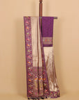 A purple and beige silk saree draped over a wooden hanger against a solid beige background. The saree features a rich purple border with intricate multicolor embroidery depicting birds and floral motifs, paired with a shimmering beige body and a matching purple dupatta.