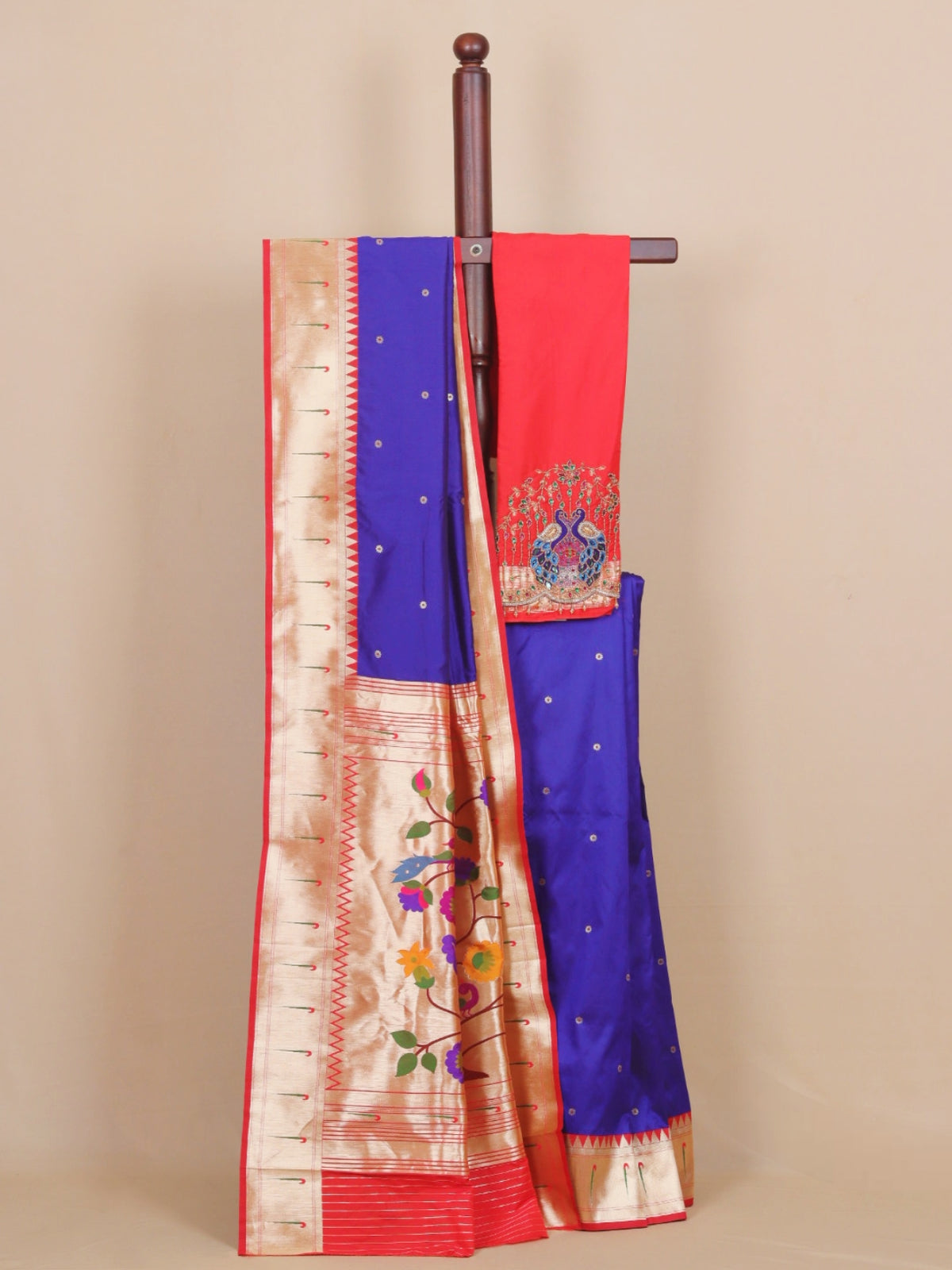 A royal blue and red saree draped over a wooden hanger against a beige background. The saree features a wide gold zari border with floral embroidery, a red pallu with peacock motifs, and small gold dots on the blue body.