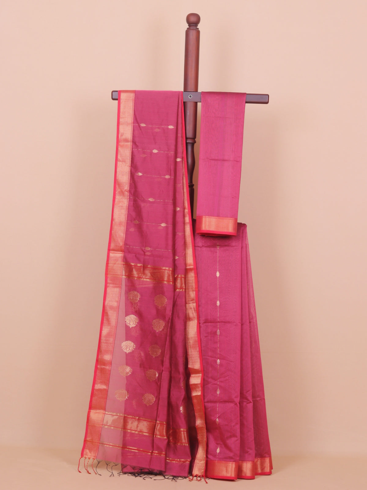 A magenta pink silk saree draped over a wooden hanger against a solid beige background. The saree features a wide gold zari border, small gold and white mirror work dots, and a matching pink blouse piece.