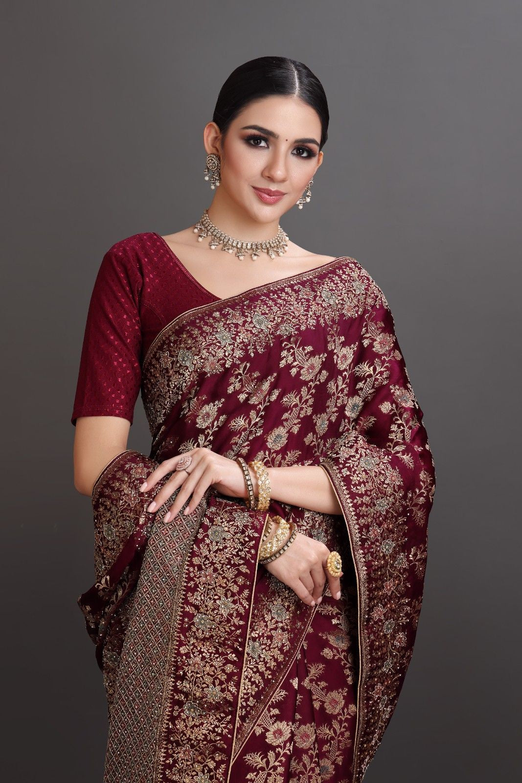 Woman wearing a maroon saree with gold patterns against a gray background