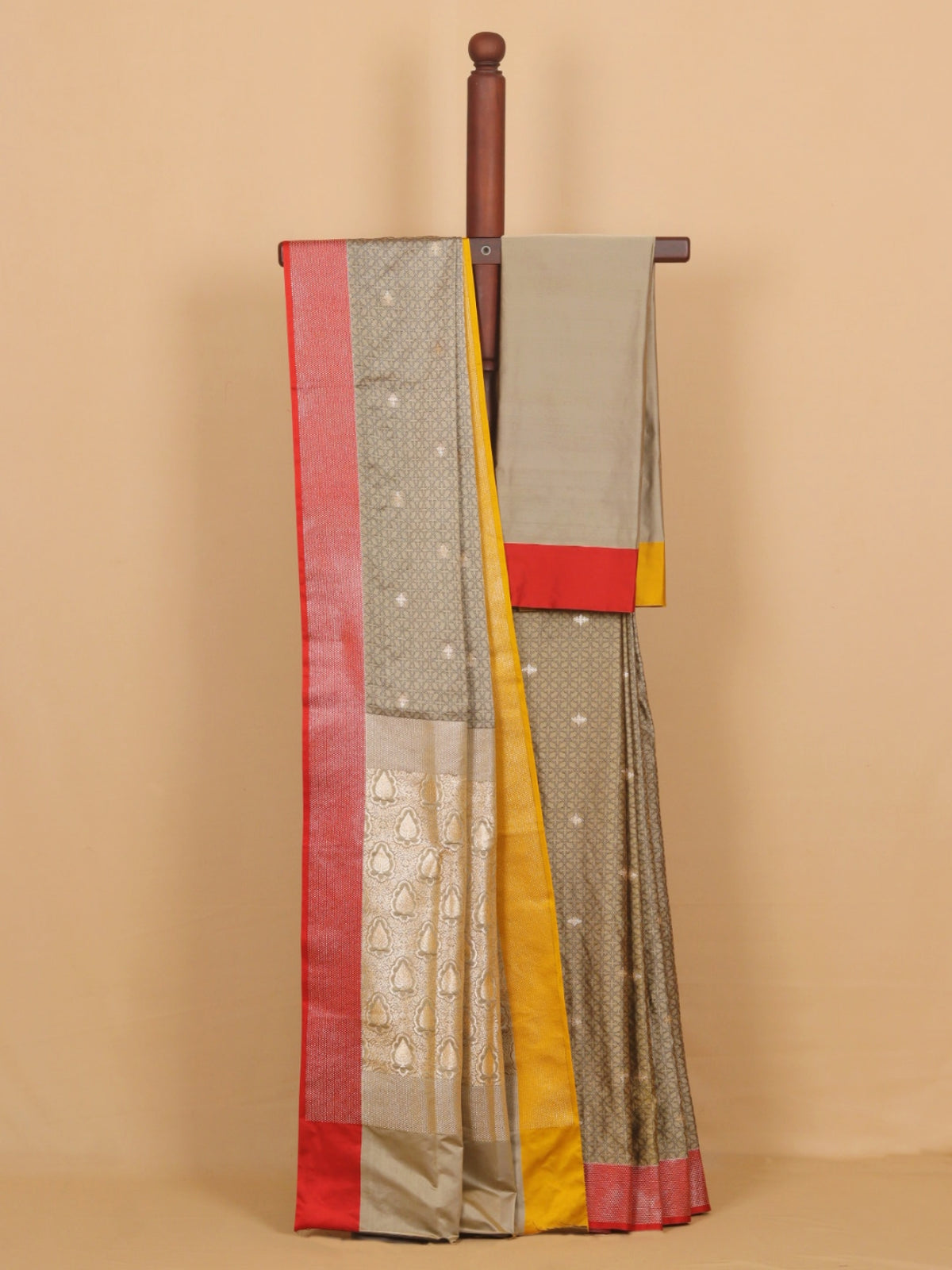 A traditional Indian saree draped over a wooden hanger against a solid beige background. The saree has a gray body with small white motifs, a red border, a yellow border, and a light gray pallu with a red and yellow border.