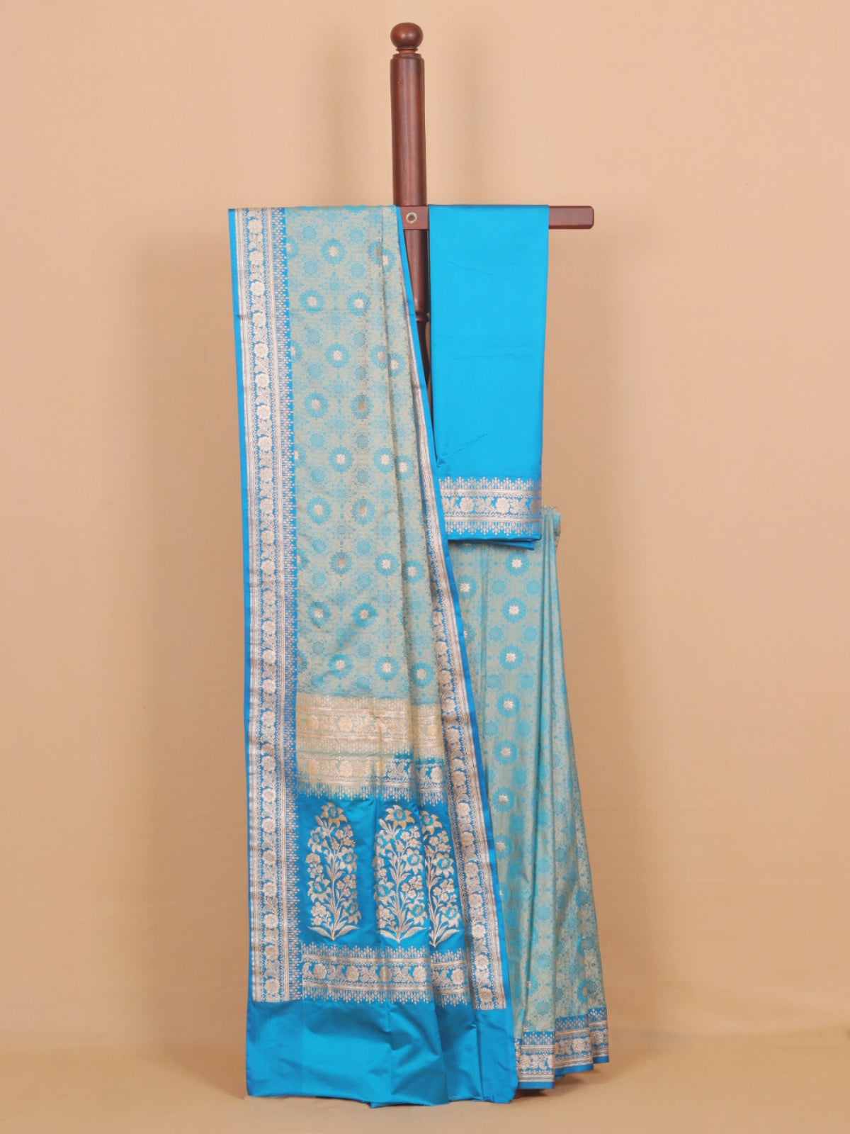 A light blue and turquoise silk saree with intricate silver zari motifs, draped over a wooden hanger against a solid beige background.