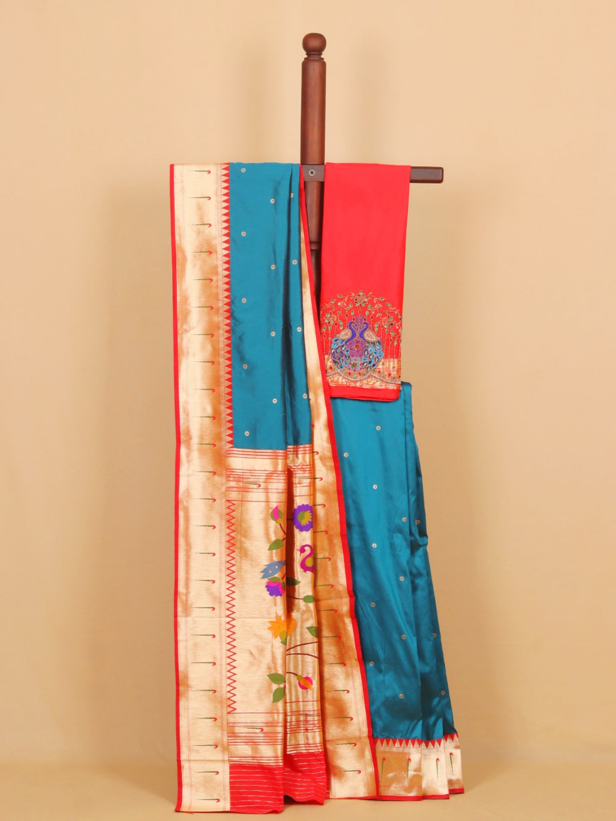 A traditional Indian silk saree draped over a wooden hanger against a solid beige background. The saree features a vibrant teal blue body with small gold motifs, a red pallu with a peacock embroidery, and a wide beige zari border with gold floral patterns.