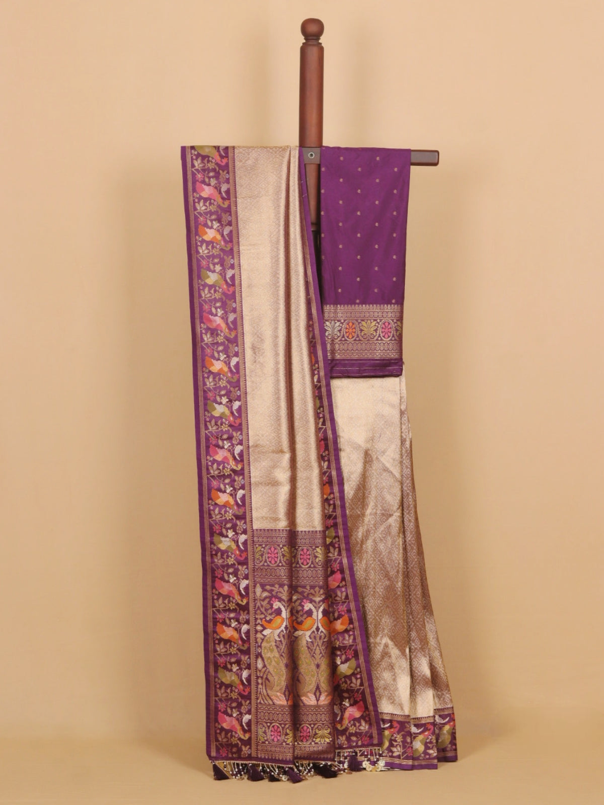 A purple and beige silk saree draped over a wooden hanger against a solid beige background. The saree features a rich purple border with intricate multicolor embroidery depicting birds and floral motifs, paired with a shimmering beige body and a matching purple dupatta.