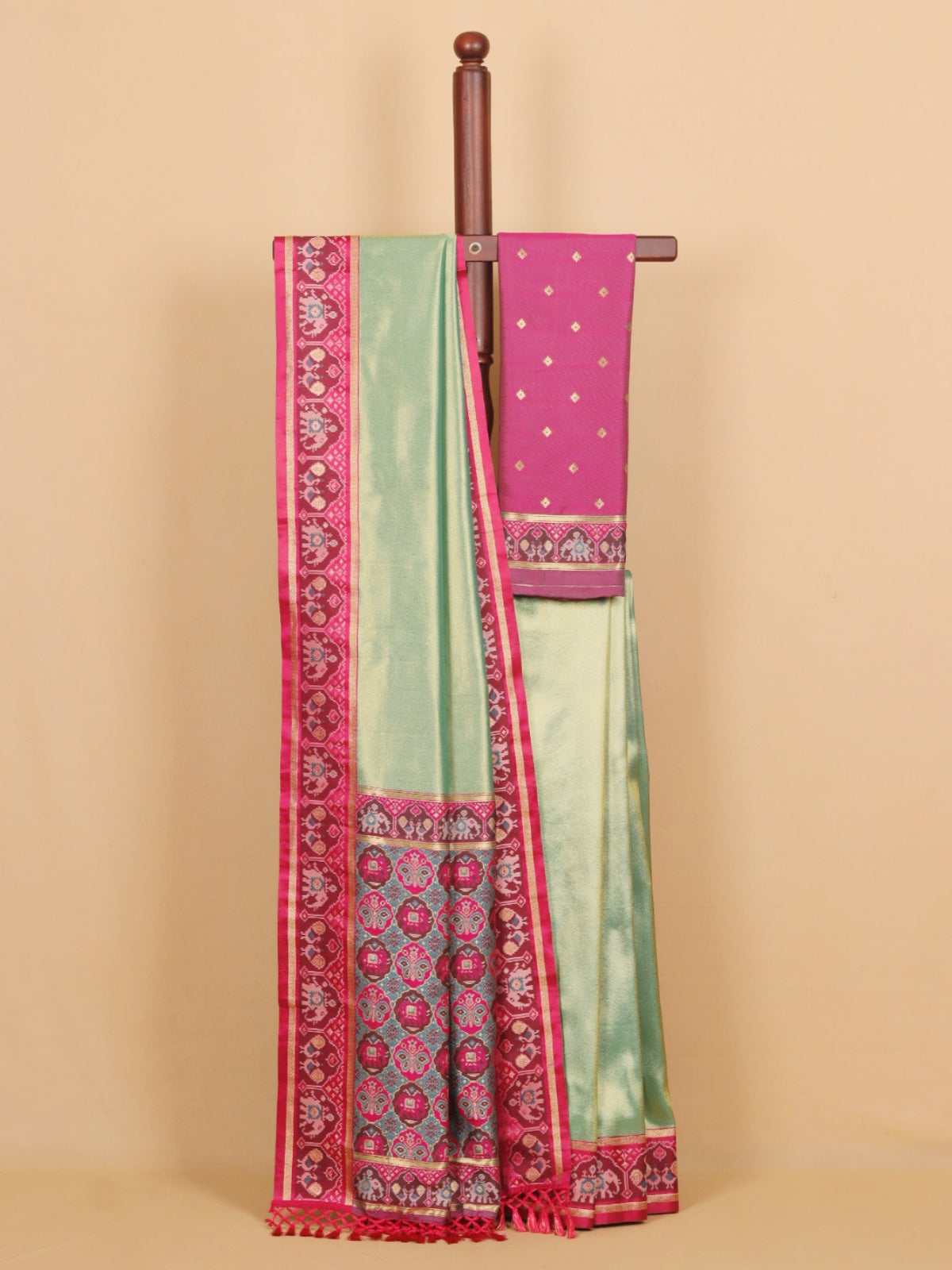 A mint green and magenta pink traditional Indian saree displayed on a wooden hanger against a solid beige background. The saree features a wide magenta border with intricate gold and white patterns, a solid mint green body, and a matching pink blouse piece with small gold dots.