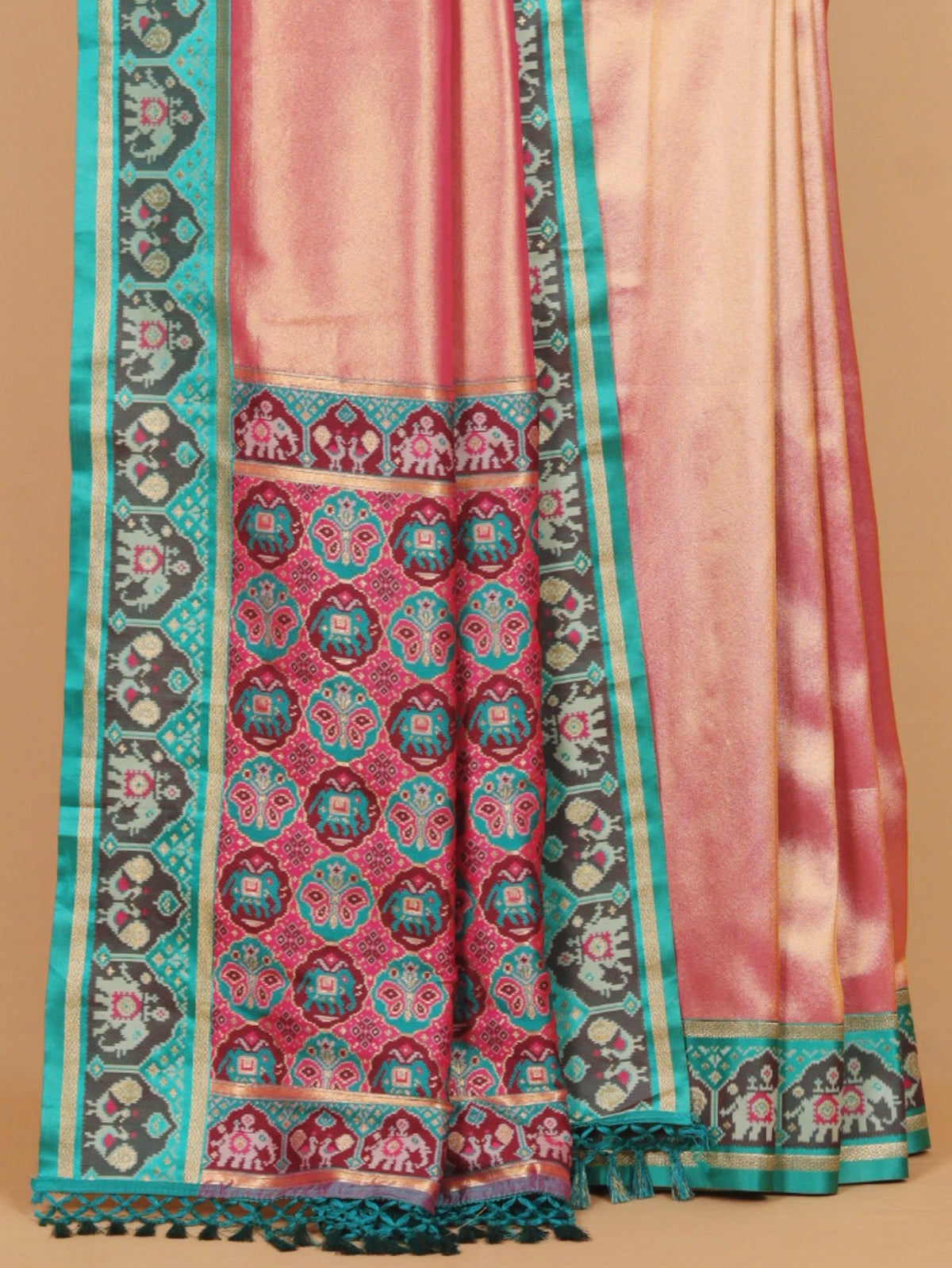 A close-up of a pink and teal patterned fabric, likely a saree or scarf, featuring intricate geometric and floral motifs with tassels on a solid tan background.