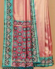 A close-up of a pink and teal patterned fabric, likely a saree or scarf, featuring intricate geometric and floral motifs with tassels on a solid tan background.