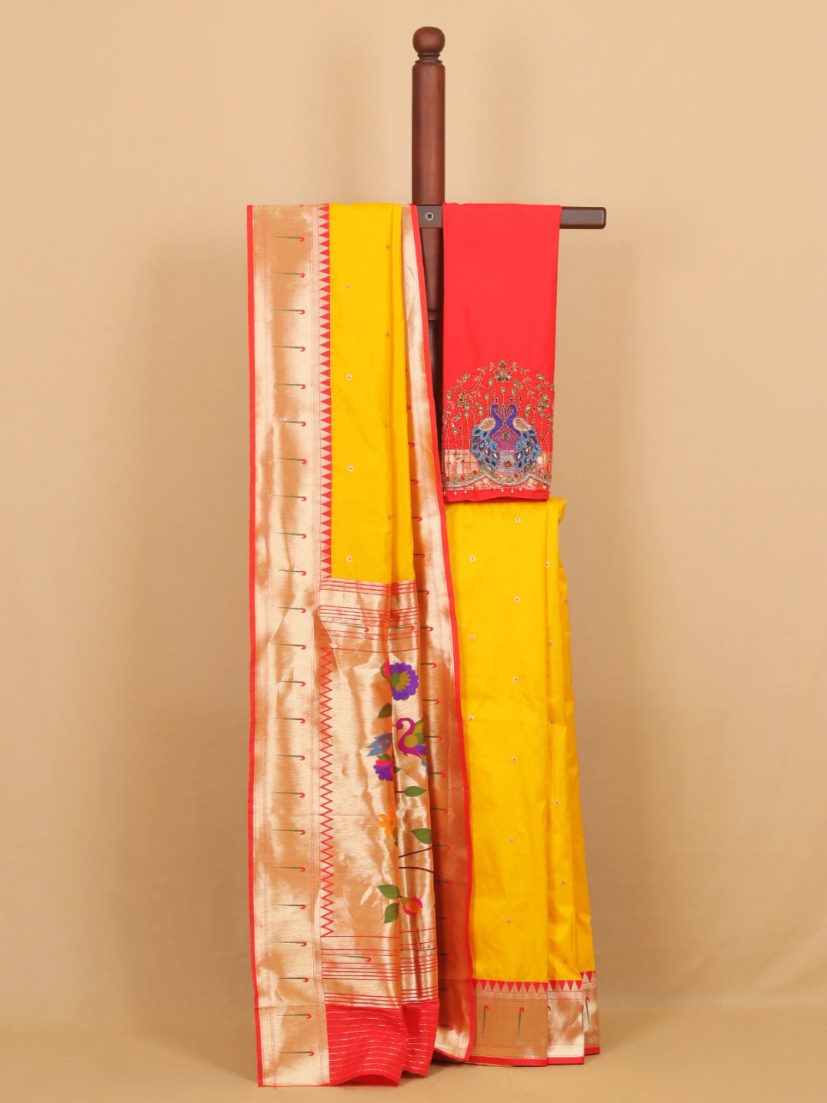 A yellow and red silk saree draped over a wooden stand against a beige background. The saree features a yellow body, a red border, and a beige zari brocade pattern with peacock embroidery on the pallu.