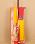 A yellow and red silk saree draped over a wooden stand against a beige background. The saree features a yellow body, a red border, and a beige zari brocade pattern with peacock embroidery on the pallu.