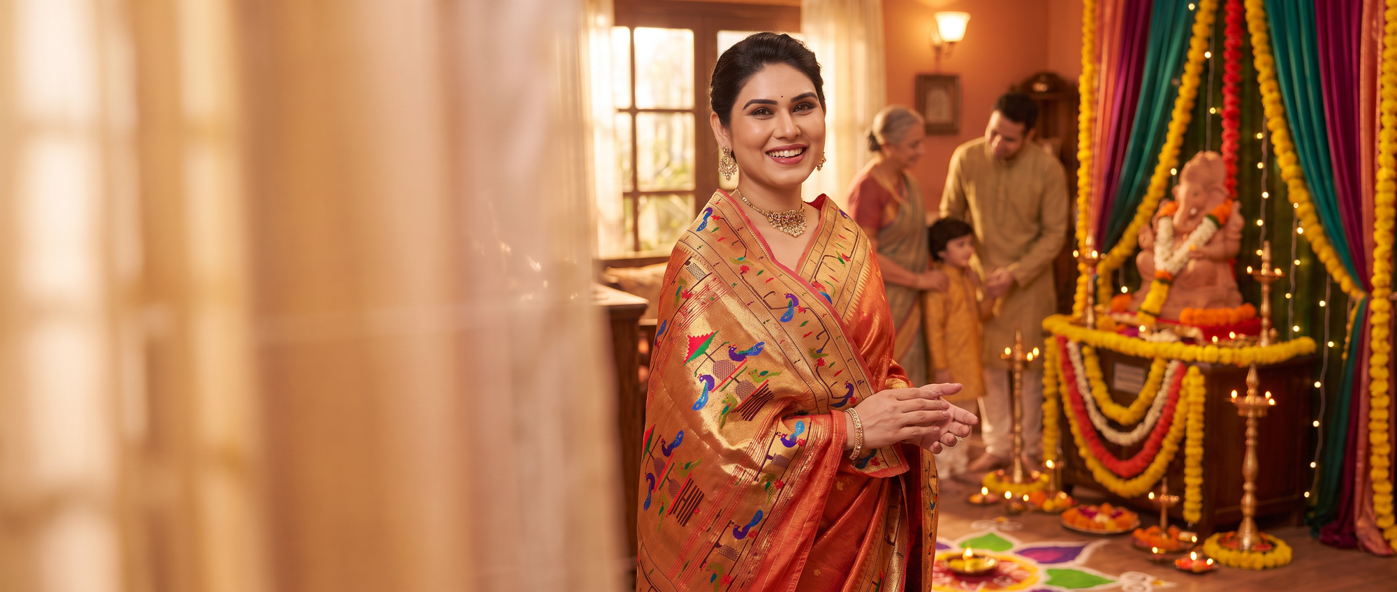 A woman in a golden-orange silk saree with colorful embroidery stands in a decorated indoor setting, smiling and holding a glass. The background features warm lighting, traditional decor, and other people in soft-focus attire.