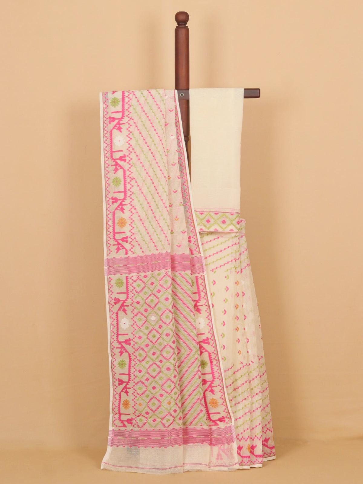 A cream-colored cotton saree with pink and green geometric patterns and a matching cream blouse, draped over a wooden hanger against a solid beige background.